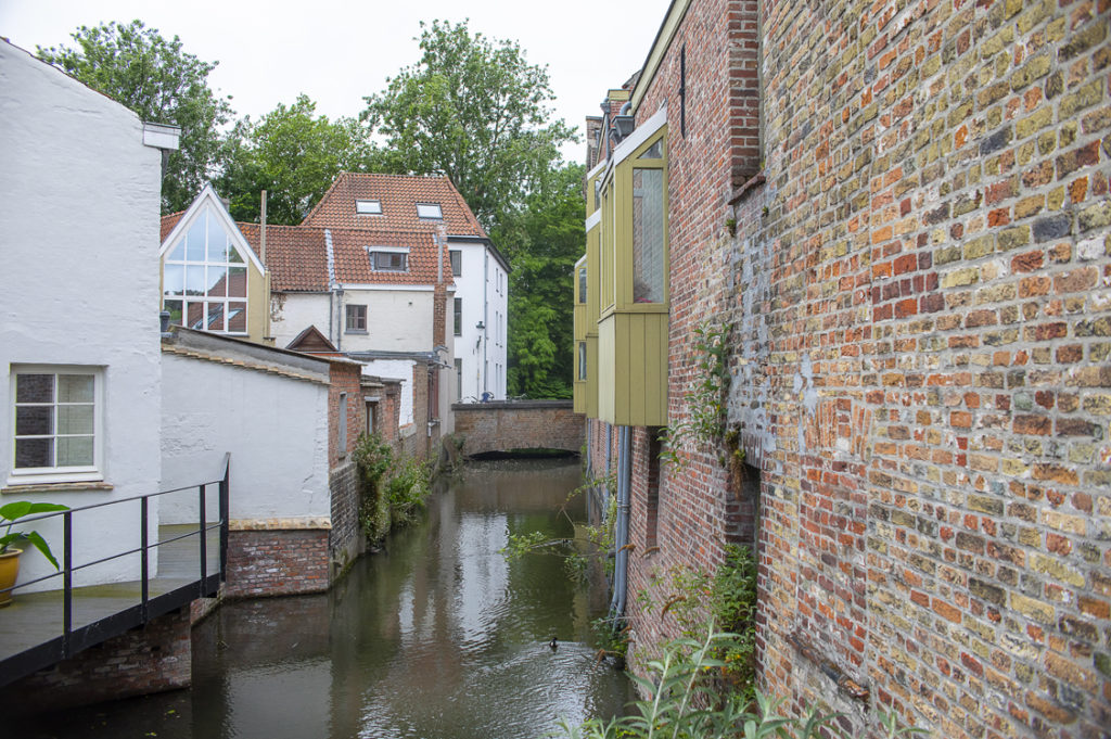 A part of a canal in the city of Bruges in Belgium. We see houses on both sides and a duck is swimming towards the end of the canal. The sky is gray, and the brick buildings are reflected in the water