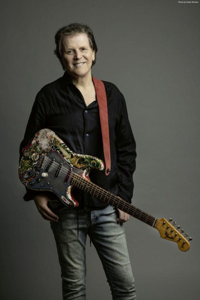 Trevor Rabin standing with his guitar hanging to his right side. He's smiling in to the camera.
