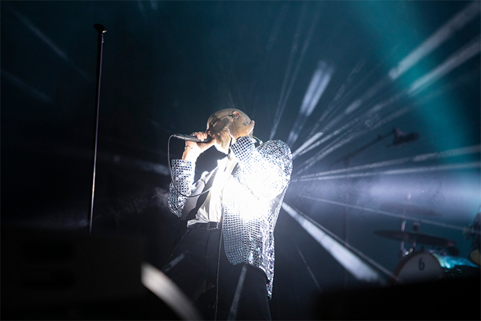 Sivert Høyem singing into a microphone while wearing a mirror ball jacket reflecting the light all over the place.